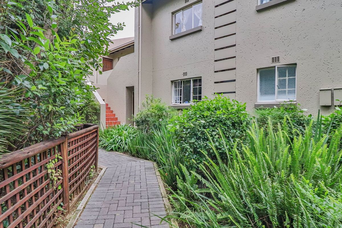 A view of the apartment building exterior featuring a paved walkway, landscaped garden area, and a timber lattice fence.