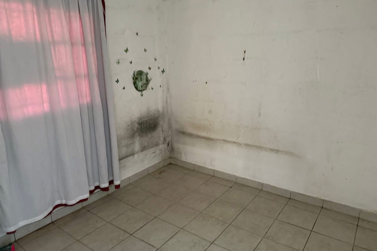 An empty bedroom featuring tiled flooring and a curtain rail, currently showing significant signs of wall dampness and surface deterioration requiring maintenance.
