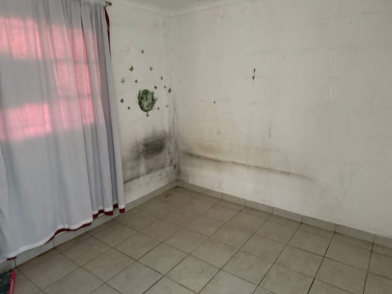 An empty bedroom featuring tiled flooring and a curtain rail, currently showing significant signs of wall dampness and surface deterioration requiring maintenance.