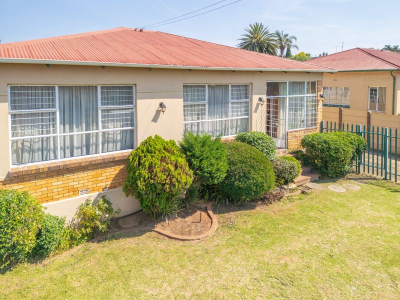 Front elevation of a single-story house featuring a corrugated metal roof, brick base detailing, and established garden landscaping with perimeter security fencing.