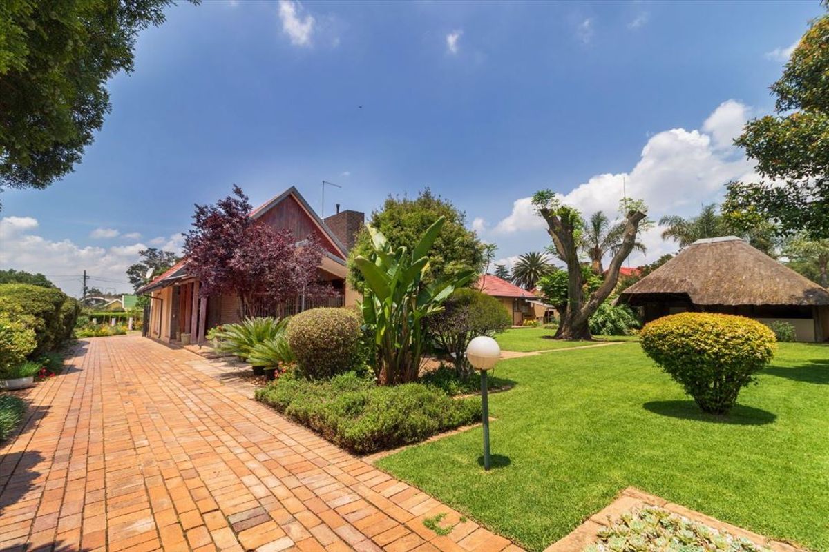 A well-maintained rear garden featuring a brick-paved pathway, lush lawn, mature landscaping, and a traditional thatched lapa structure.