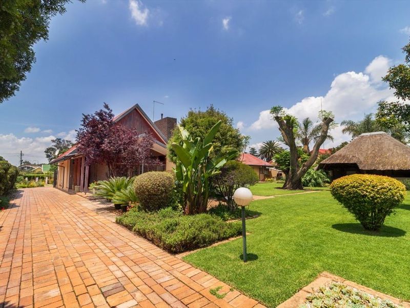 A well-maintained rear garden featuring a brick-paved pathway, lush lawn, mature landscaping, and a traditional thatched lapa structure.