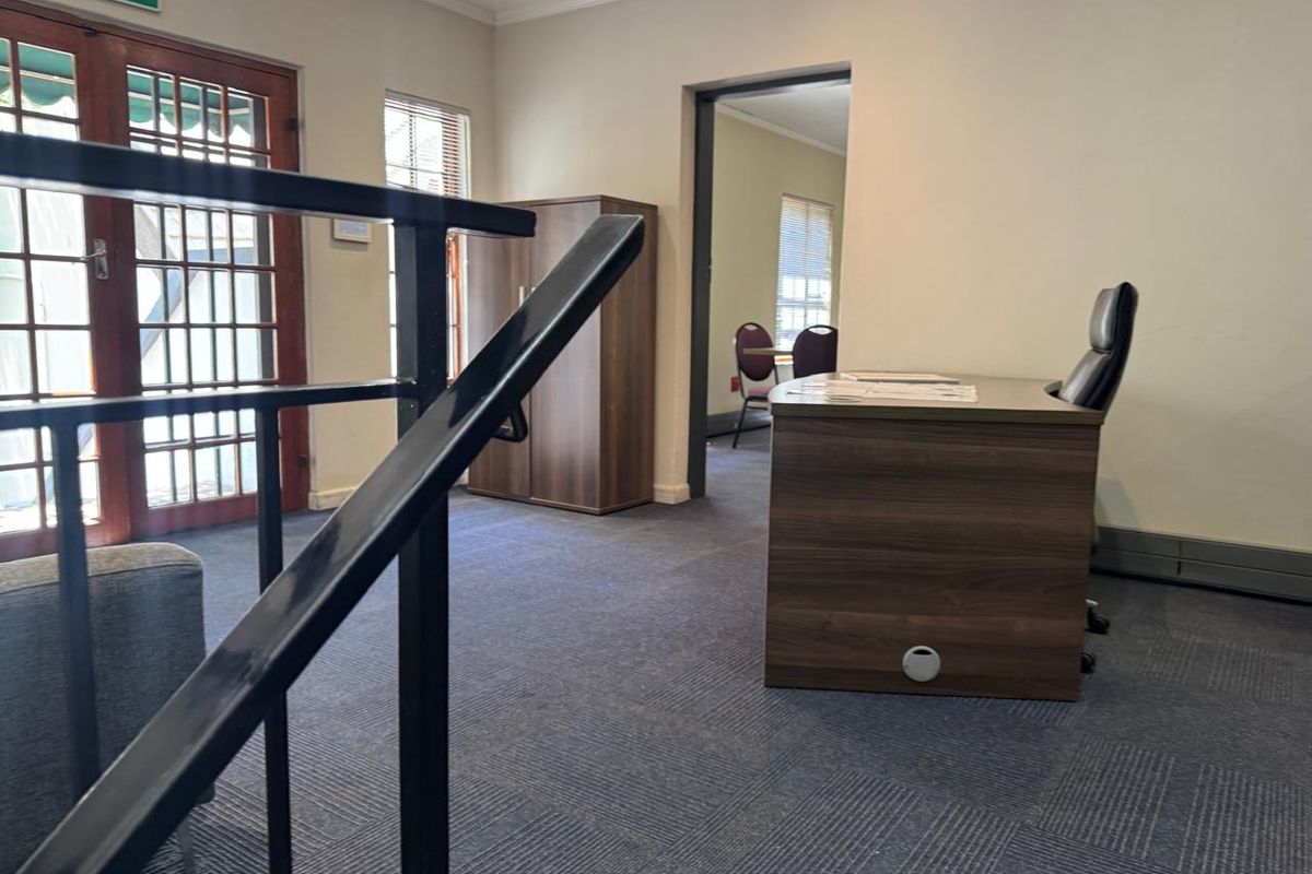 A functional office space featuring carpeted flooring, a metal staircase railing, and access to an adjacent room through a doorway.