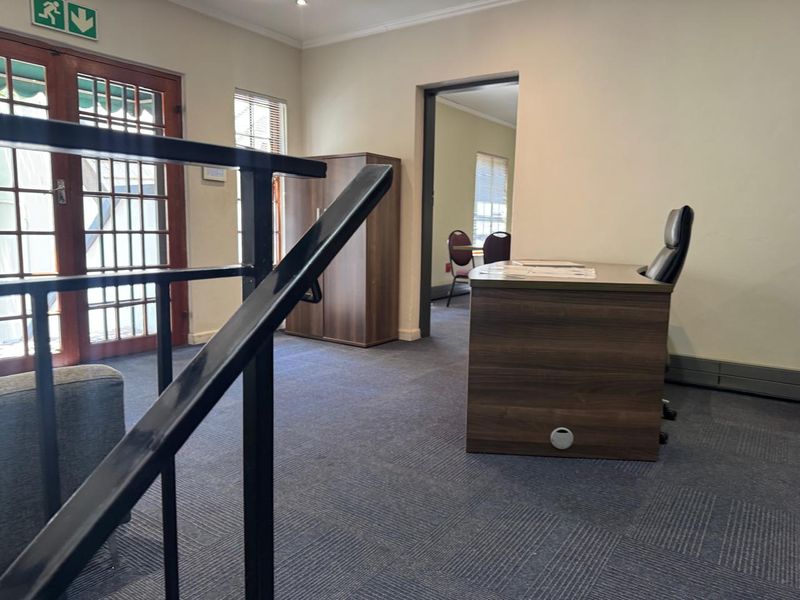A functional office space featuring carpeted flooring, a metal staircase railing, and access to an adjacent room through a doorway.