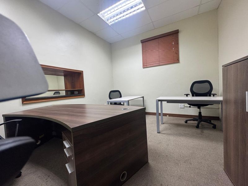 A functional office space featuring carpeted flooring, a suspended ceiling with integrated lighting, and an internal window with wooden blinds.