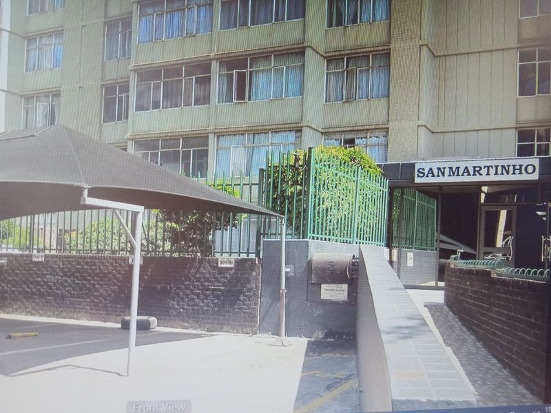 Front exterior view of the San Martinho apartment complex, featuring a paved driveway, shade cloth carport, and secure boundary walling.