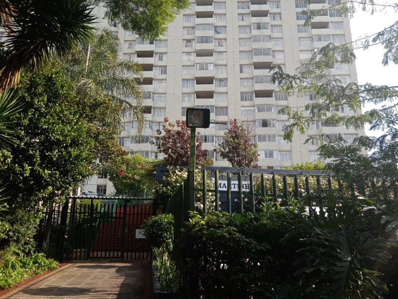 Exterior view of a high-rise apartment building featuring a secure gated entrance, paved walkway, and established garden landscaping.