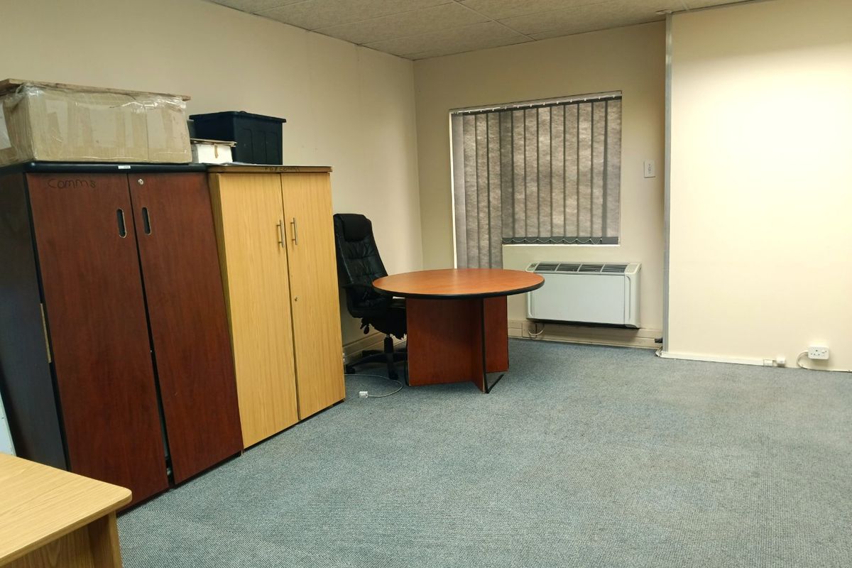 A functional office space featuring carpeted flooring, a suspended ceiling, and a wall-mounted air conditioning unit with vertical window blinds.