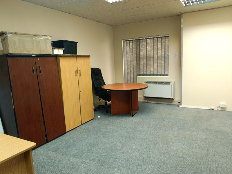 A functional office space featuring carpeted flooring, a suspended ceiling, and a wall-mounted air conditioning unit with vertical window blinds.