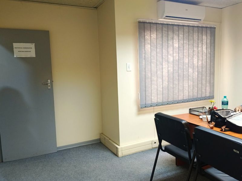 A functional office space featuring carpeted flooring, a wall-mounted air conditioning unit, and vertical window blinds.