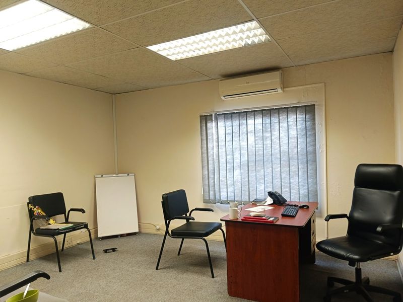 A functional office space featuring a suspended ceiling, recessed lighting, wall-mounted air conditioning, and vertical window blinds.