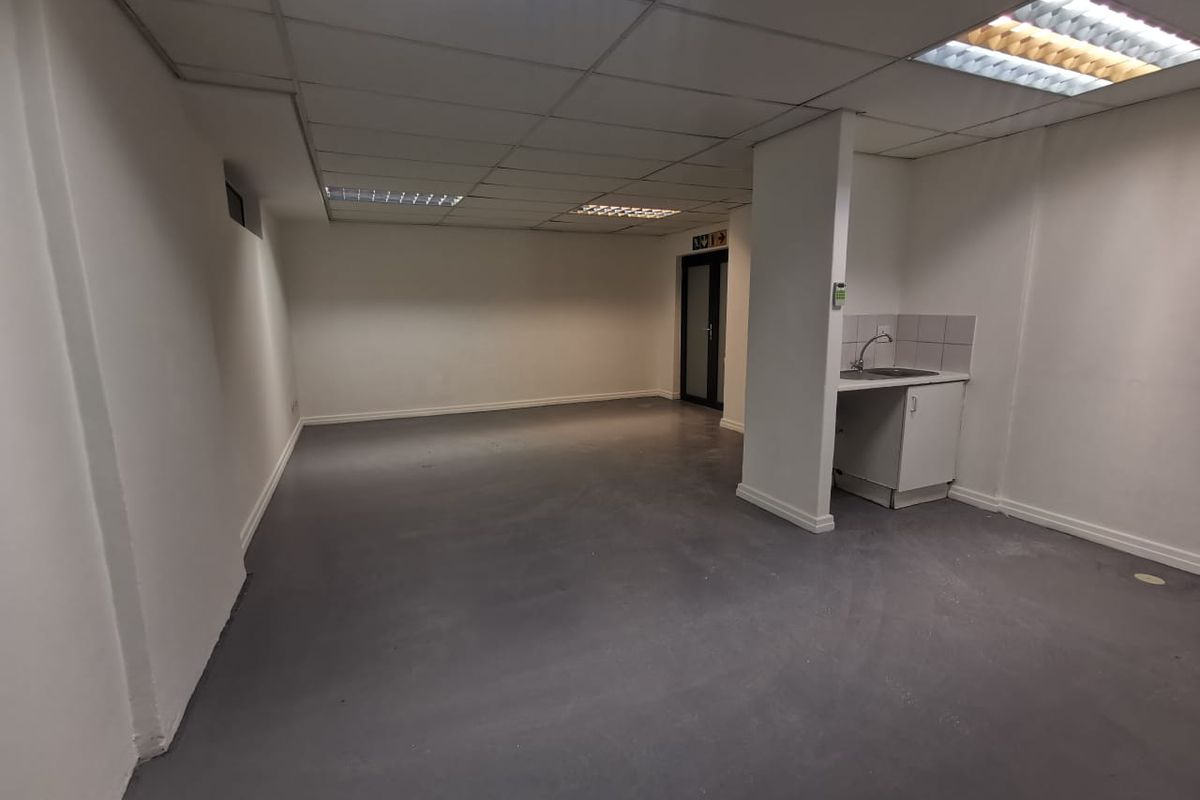 An open-plan commercial office space featuring a suspended ceiling, recessed lighting, painted concrete floors, and a small kitchenette area with a sink.