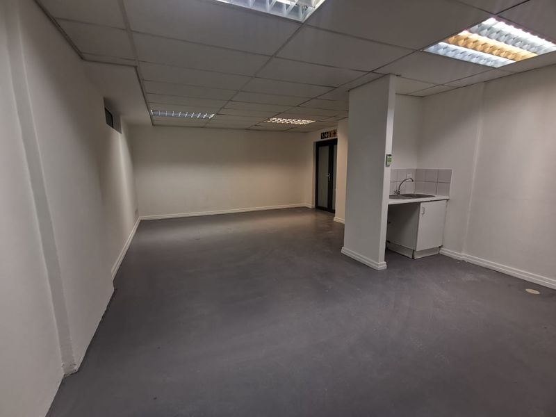 An open-plan commercial office space featuring a suspended ceiling, recessed lighting, painted concrete floors, and a small kitchenette area with a sink.