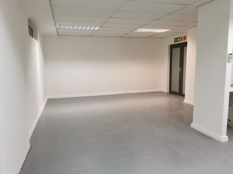 A clean, vacant commercial office space featuring a suspended ceiling, recessed lighting, and durable painted concrete flooring with an emergency exit.