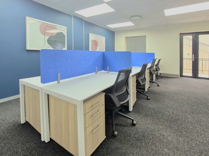 A modern commercial office space featuring built-in workstation desks with integrated storage, acoustic privacy screens, carpeted flooring, and recessed ceiling lighting.