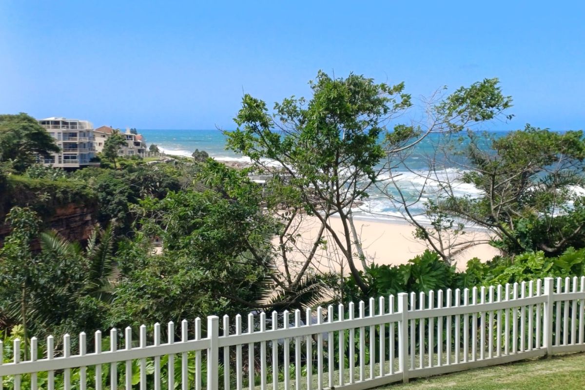 Scenic coastal view overlooking the beach and ocean, framed by a white picket fence and lush greenery.
