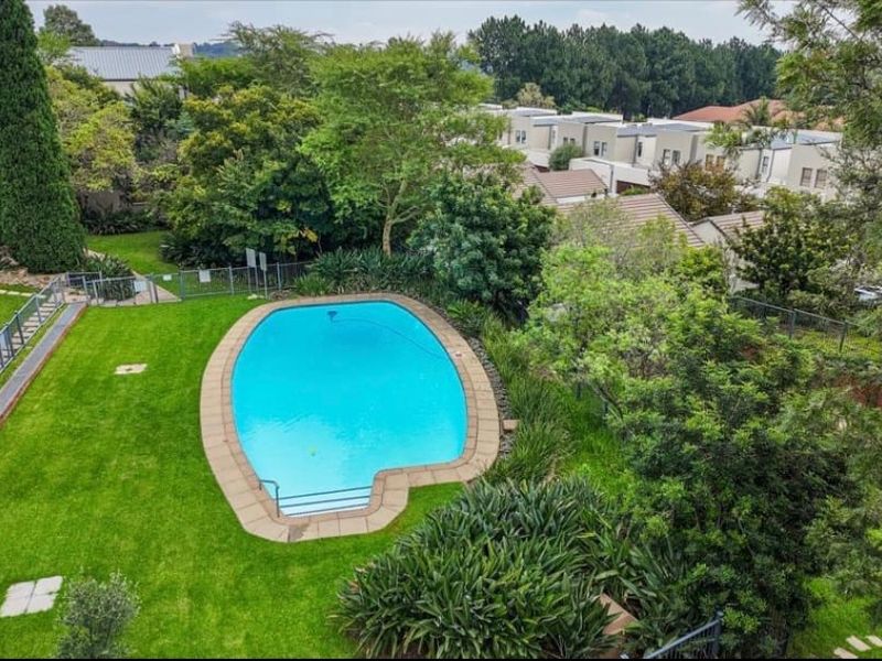 Aerial view of a well-maintained communal swimming pool area surrounded by lush green lawns and established garden landscaping within the apartment complex.