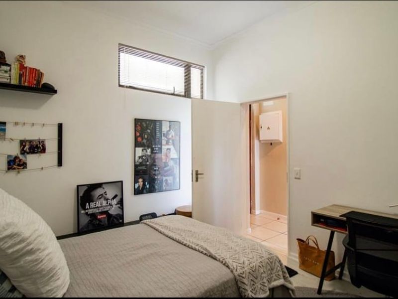 A well-lit bedroom featuring neutral walls, a horizontal window with blinds, and direct access to an adjoining hallway.
