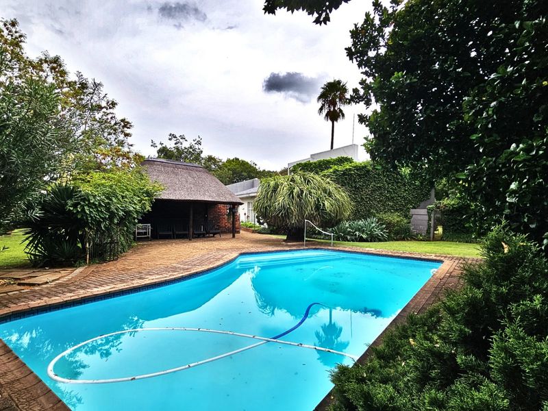A well-maintained outdoor pool area featuring a swimming pool, brick-paved surround, and a traditional thatched lapa set within a lush, landscaped garden.