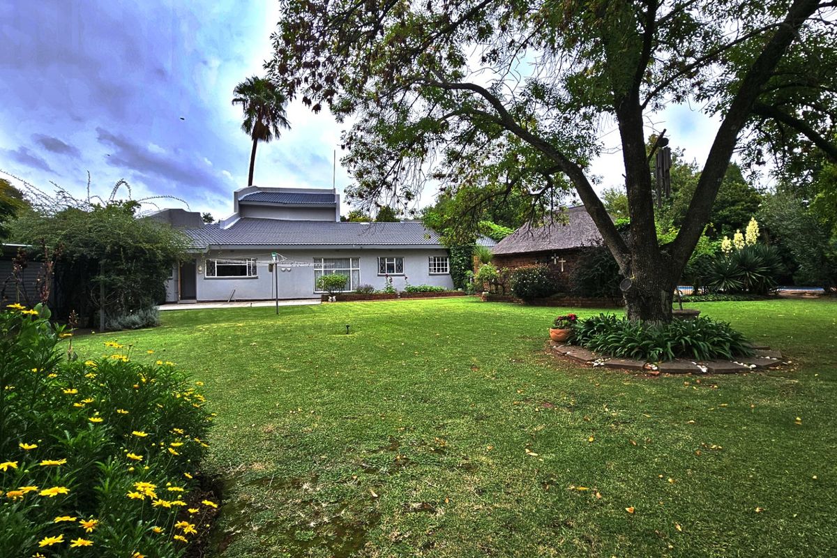 A spacious rear garden featuring a well-maintained lawn, mature trees, and a view of the main house with a tiled roof and a separate thatched structure.