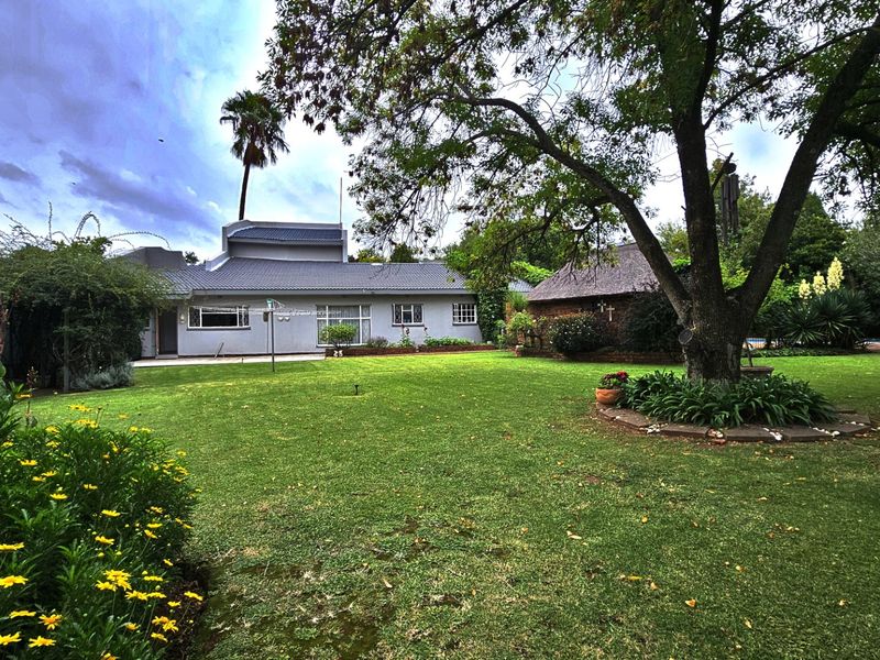 A spacious rear garden featuring a well-maintained lawn, mature trees, and a view of the main house with a tiled roof and a separate thatched structure.