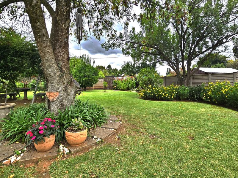 A well-maintained, spacious garden featuring mature trees, lush green lawn, and established flower beds enclosed by boundary fencing.