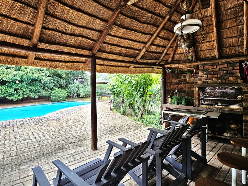 Covered outdoor patio featuring a traditional thatched roof, built-in brick braai, and direct access to a swimming pool and paved garden area.