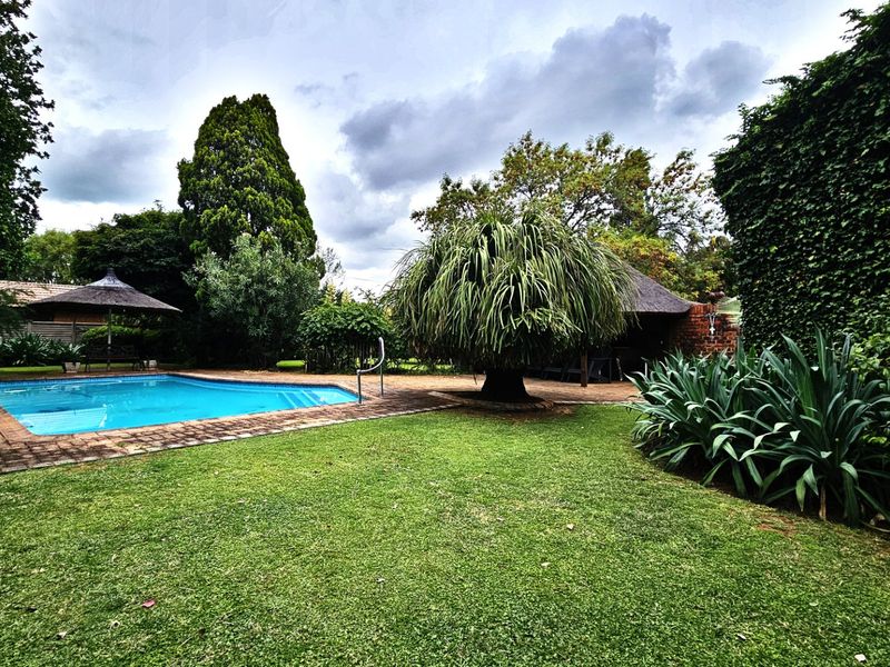A well-maintained outdoor pool area featuring a swimming pool with brick coping, surrounded by a lush garden and a traditional thatched lapa.