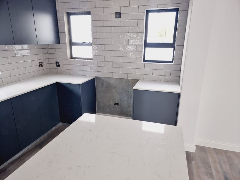 Modern kitchen featuring dark cabinetry, white stone countertops, and grey subway tile splashbacks. The space is currently awaiting final appliance installation.