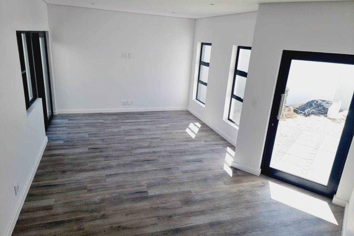 A bright, empty living room featuring modern wood-look laminate flooring, recessed ceiling lighting, and large black-framed windows and doors leading to the exterior.