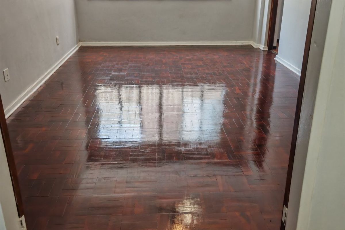 A bright, empty living room featuring polished dark parquet flooring, neutral walls, and large windows fitted with security bars.