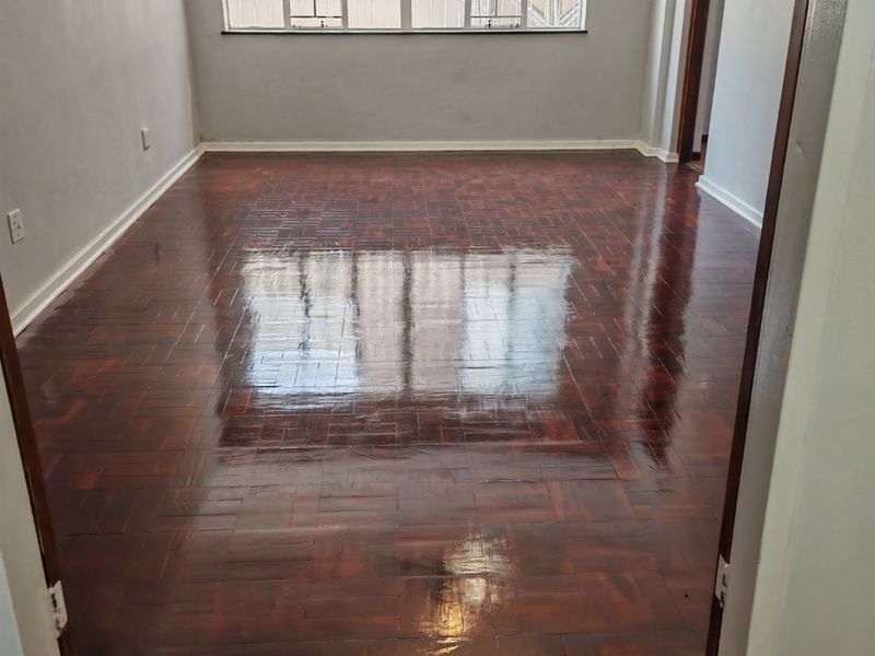 A bright, empty living room featuring polished dark parquet flooring, neutral walls, and large windows fitted with security bars.