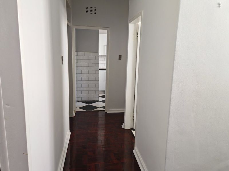 A clean, empty hallway featuring dark parquet flooring and white walls, providing access to adjacent rooms including a kitchen with tiled splashbacks.