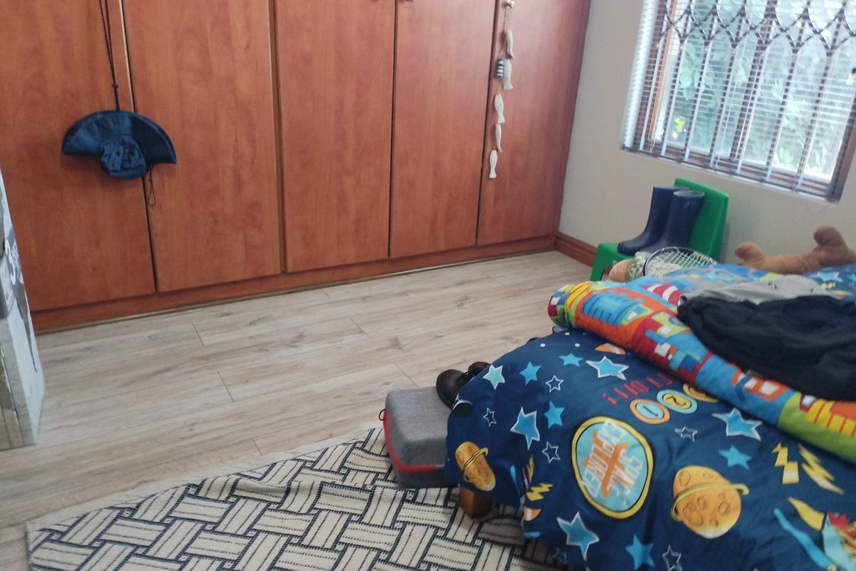 A bedroom featuring laminate flooring, built-in wooden cupboards, and a window fitted with security bars and venetian blinds.