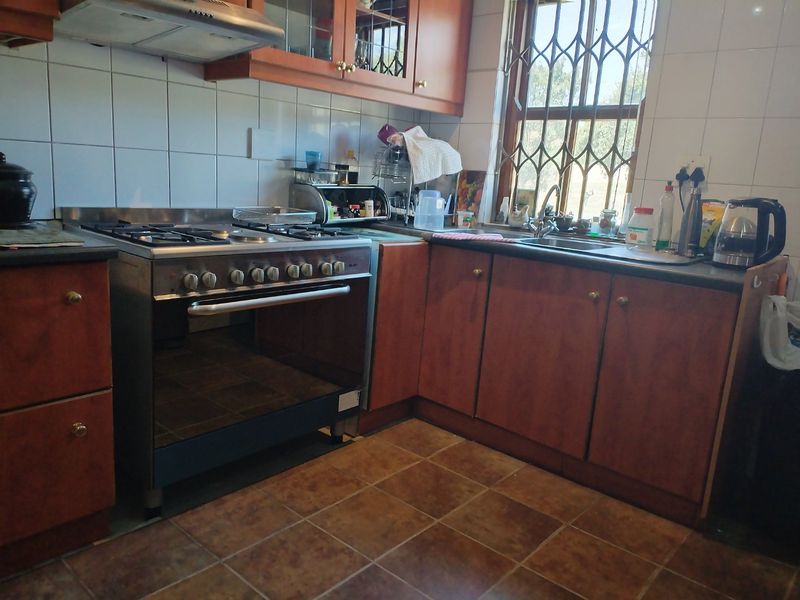 A functional kitchen featuring wooden cabinetry, a gas stove with extractor fan, tiled backsplash, and a double sink with a security-gated window.
