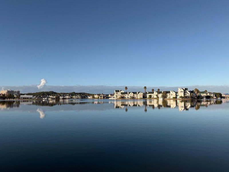 A scenic wide-angle view of a residential apartment complex situated along a calm, reflective waterfront under a clear blue sky.