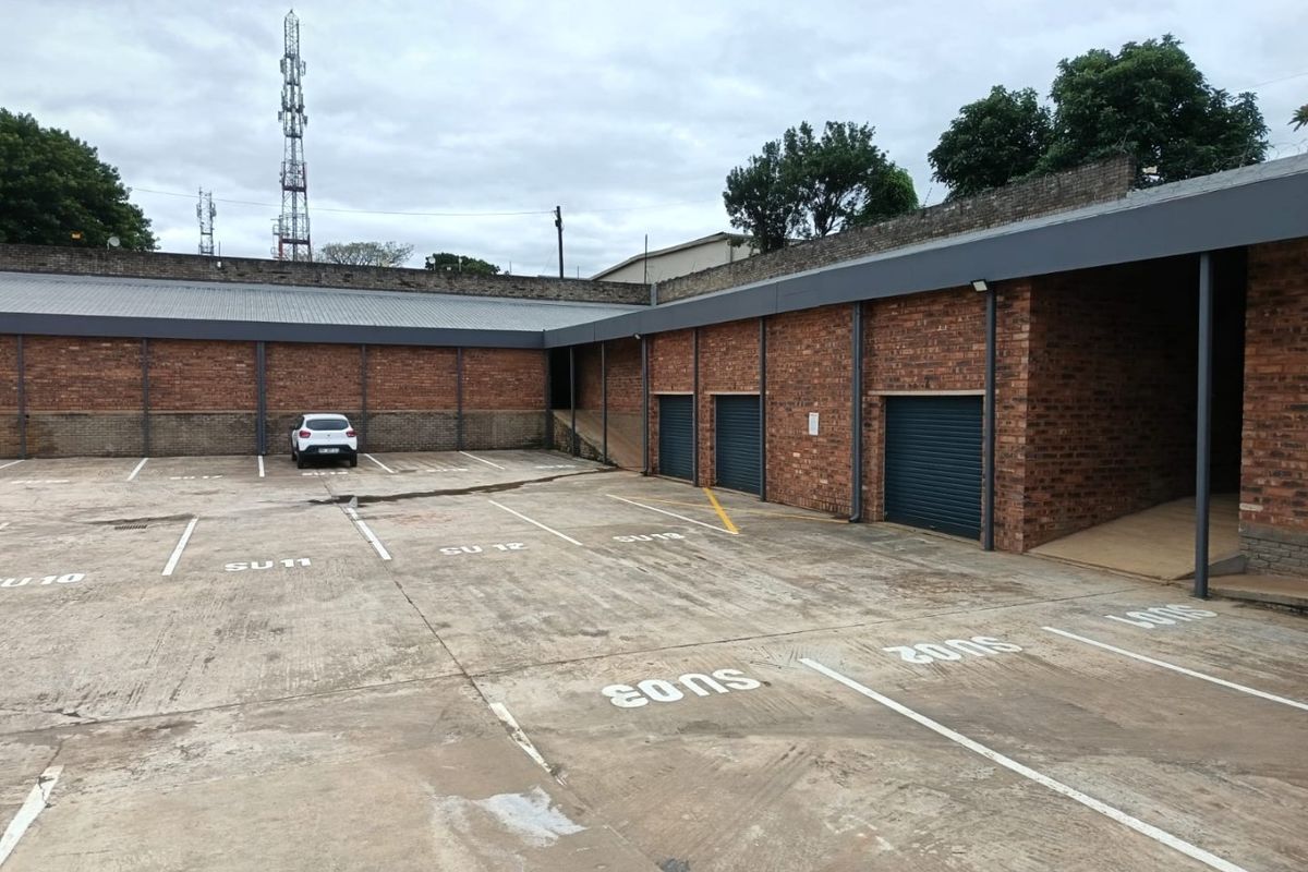 Commercial rear exterior area featuring a paved parking lot with marked bays, brick perimeter walls, and multiple metal roller shutter access doors.