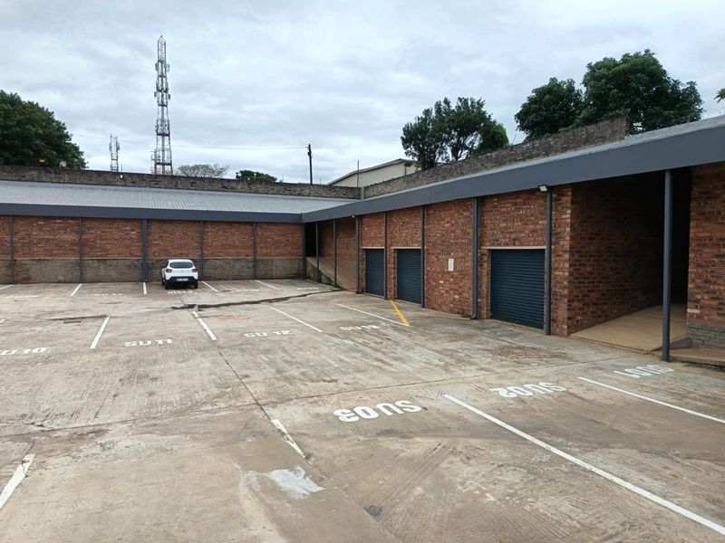 Commercial rear exterior area featuring a paved parking lot with marked bays, brick perimeter walls, and multiple metal roller shutter access doors.