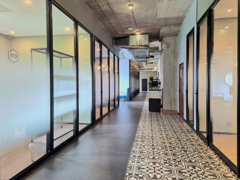 Modern commercial hallway featuring exposed concrete ceilings, patterned floor tiles, and glass-partitioned office suites with black metal frames.