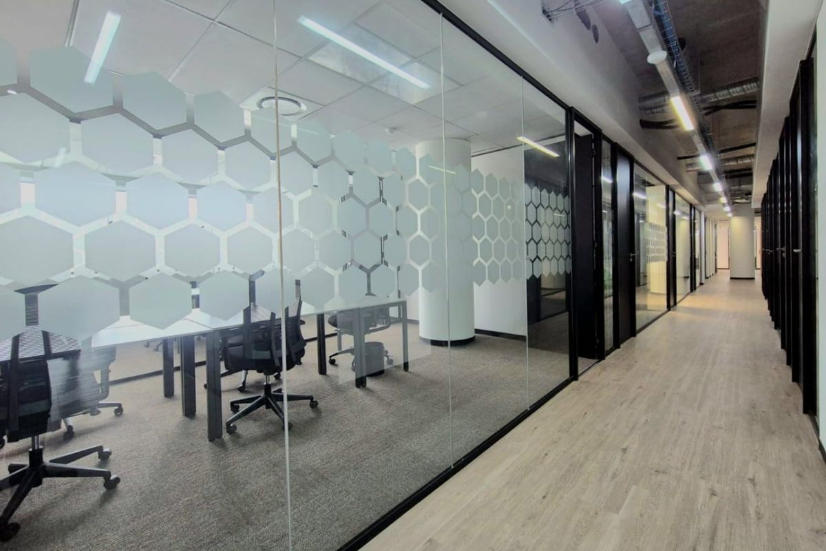Modern commercial office hallway featuring glass partitions with geometric privacy film, exposed industrial ceilings, and a mix of laminate and carpeted flooring.