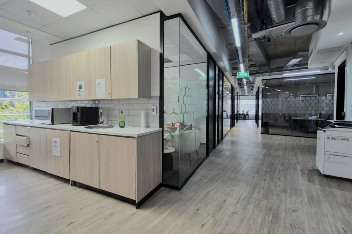 Modern commercial kitchenette featuring light wood cabinetry, sink, and glass-partitioned office spaces with an industrial-style exposed ceiling.