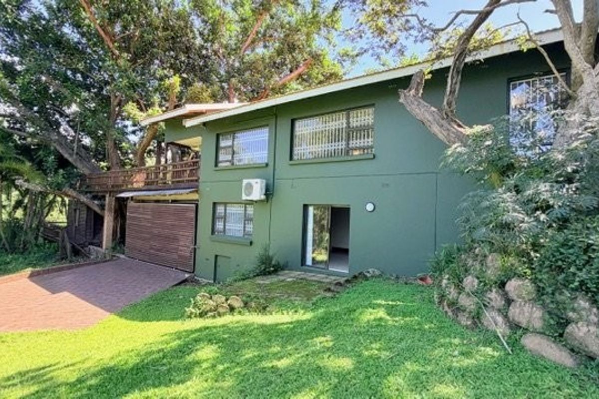 Rear view of the property featuring a timber deck, paved driveway, and established garden surroundings with mature trees.