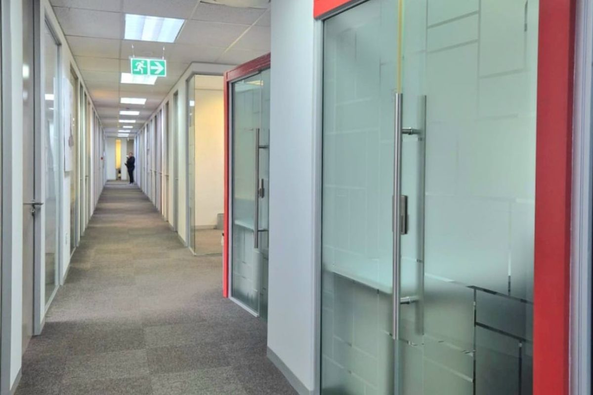 A commercial office hallway featuring carpeted flooring, suspended ceilings with integrated lighting, and modern glass partition walls with red framing.