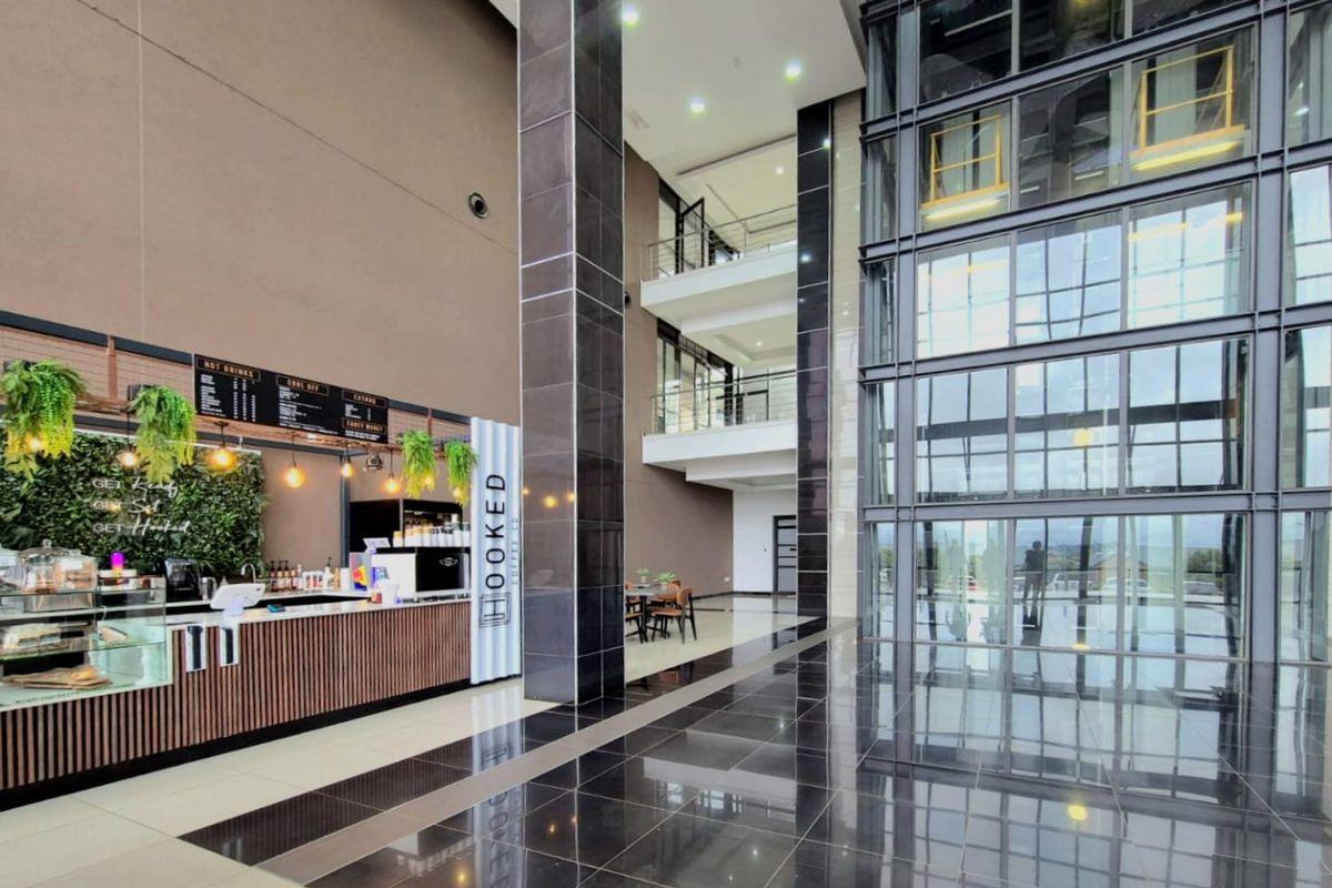 Modern commercial atrium featuring a high-ceiling glass facade, polished tiled flooring, and a built-in coffee service counter within a multi-level open-plan lobby.