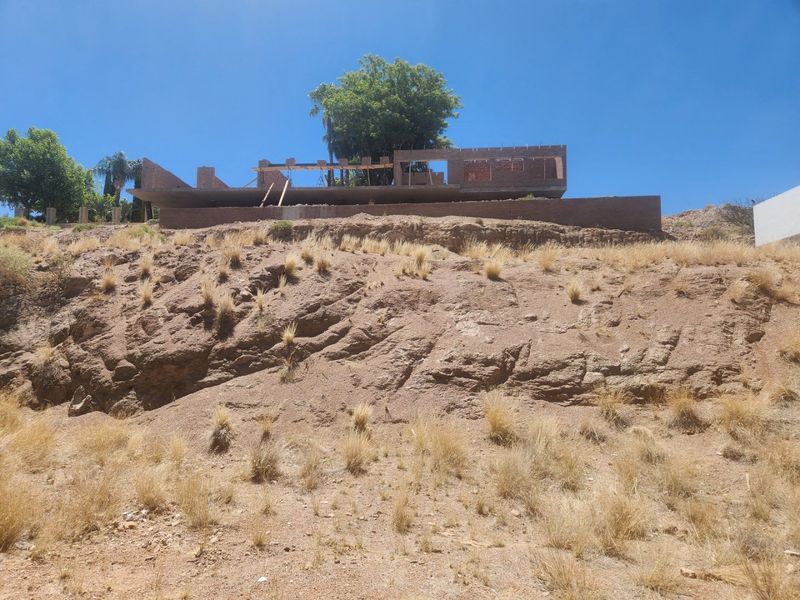 A view of a residential property under construction, featuring a brick structure and retaining wall on a sloped, undeveloped plot.