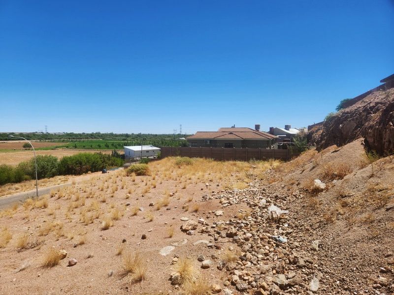 An elevated view of an unimproved residential plot featuring rocky terrain, natural vegetation, and a surrounding precast concrete boundary wall.