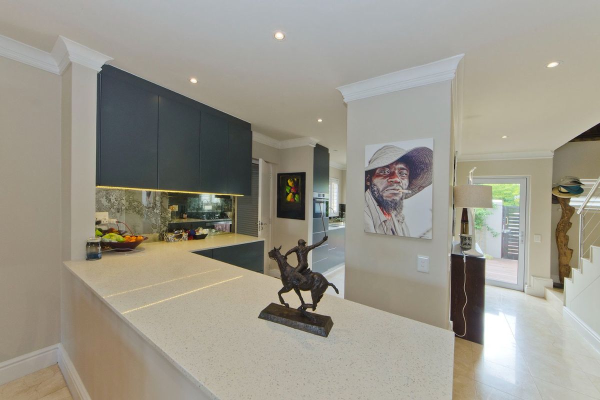 Modern kitchen featuring sleek dark cabinetry, stone countertops, a mirrored splashback, and recessed lighting, seamlessly flowing into the adjacent living area.