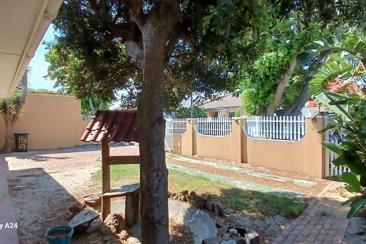 A spacious rear garden area featuring a paved walkway, perimeter boundary wall with white fencing, and mature trees providing shade.