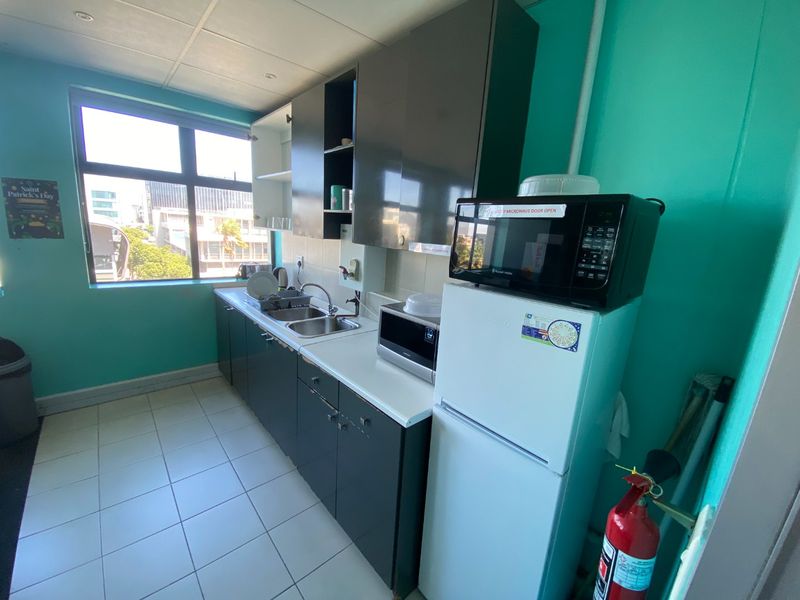 A functional commercial kitchenette featuring dark cabinetry, a stainless steel sink, tiled flooring, and a large window providing natural light.