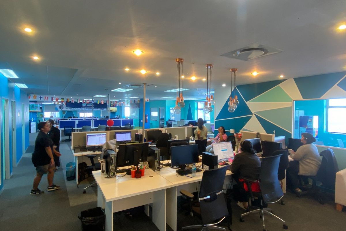 A spacious open-plan commercial office area featuring built-in workstations, modern pendant lighting, recessed ceiling lights, and carpeted flooring.